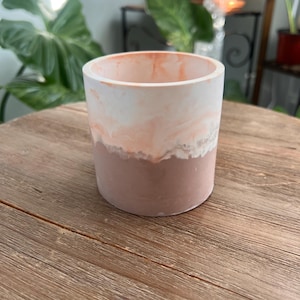 May include: A cylindrical planter with a two-tone design. The bottom half is a muted mauve color, while the top half features a marbled effect of white and peach. The planter is sitting on a wooden surface.