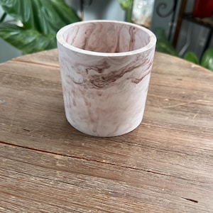May include: A cylindrical planter with a marbled design in shades of white and brown. The planter is made of a solid material and sits on a wooden surface. The interior of the planter is visible, showcasing the same marbled pattern.