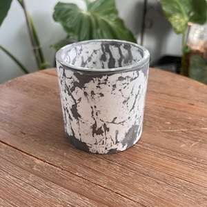 May include: A cylindrical concrete planter with a white base and dark gray splatter design. The rim and base are dark gray. The planter sits on a wooden surface. The planter is suitable for small plants and succulents.