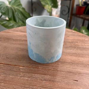 May include: A cylindrical concrete planter with a marbled blue and white design. The planter has a smooth, rounded rim and a solid base. The blue color is concentrated at the bottom, creating a watercolor effect. The planter is suitable for small plants.