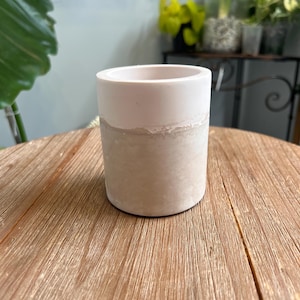May include: A cylindrical, two-toned planter. The top portion is a light pink, while the bottom is a textured, light beige. The planter is sitting on a wooden surface. The planter is suitable for small plants and succulents.