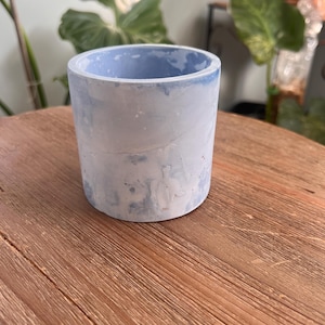 May include: A cylindrical concrete planter with a light blue interior and a marbled, light blue and white exterior. The planter sits on a wooden surface and is suitable for small plants or succulents.