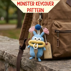 May include: A blue crocheted monster key cover with a yellow mouth, pink accents, and a blue ball on top. The key cover is attached to a backpack with a key and a luggage tag. The text "MONSTER KEYCOVER PATTERN" is at the top.