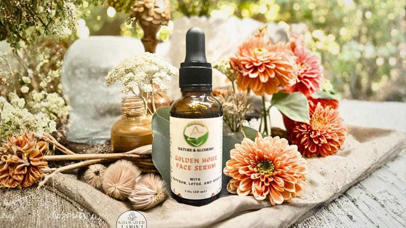 May include: A brown glass bottle of Golden Hour Face Serum with a black dropper, set against a backdrop of orange flowers and greenery. The label reads "Nature & Alchemy" and lists ingredients. The bottle is 1 fl oz (30 ml).