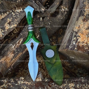 May include: A decorative dagger with a green wooden handle and a silver blade, resting on a bed of wood shavings and logs. The dagger has a matching green leather sheath with a silver emblem. The text "IGNIVAR FORGE" is visible.