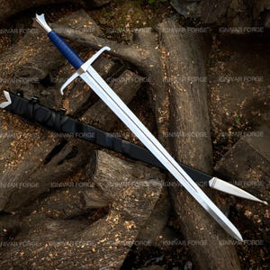 May include: A long, silver sword with a blue handle and a black sheath rests on a pile of wood. The sword has a double-edged blade and a crossguard. The sheath is made of leather. The text "IGNIVAR FORGE" is visible.
