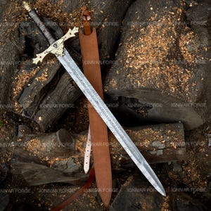May include: A detailed image of a long sword with a patterned blade and ornate gold-colored hilt. The sword rests on a bed of wood shavings and logs, with a brown leather sheath. The sword's handle is wrapped in a dark material.