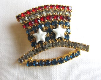 May include: A patriotic Uncle Sam hat pin, adorned with red, white, and blue rhinestones. The hat features two white star accents and a gold-toned brim, perfect for adding a touch of Americana to any outfit.