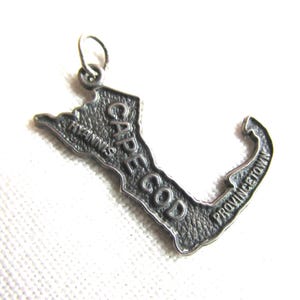 May include: A silver-colored pendant in the shape of Cape Cod, Massachusetts. The pendant has the words "CAPE COD", "HYANNIS", and "PROVINCETOWN" engraved on it. The pendant has a small loop at the top for attaching to a chain.