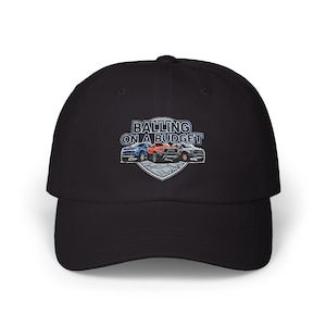 May include: Black baseball cap featuring the text "BALLING ON A BUDGET" above a graphic of three cars in blue, orange, and grey. The graphic is set on a shield-shaped background.