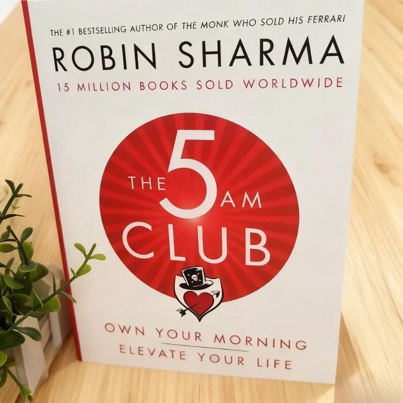 Może przedstawiać: Książka "The 5 AM Club" autorstwa Robina Sharma. Okładka przedstawia duży czerwony okrąg z białą cyfrą 5 i napisem "THE AM CLUB". Tytuł książki znajduje się nad okręgiem, a nazwisko autora u g&oacute;ry.