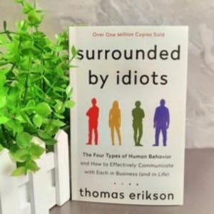 Może przedstawiać: Książka zatytułowana "surrounded by idiots" autorstwa Thomasa Eriksona. Okładka zawiera tekst "Over One Million Copies Sold" oraz grafikę czterech kolorowych postaci. Widoczny jest r&oacute;wnież podtytuł książki. Zielona roślina i białe ogrodzenie w tle.