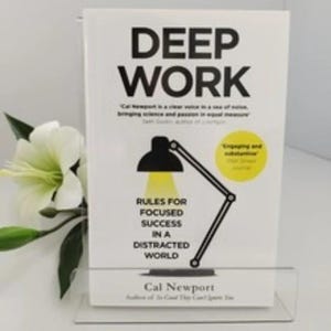 Może przedstawiać: Książka zatytułowana "DEEP WORK" z czarnym tekstem na białym tle. Okładka zawiera czarną grafikę lampy biurkowej i podtytuł "RULES FOR FOCUSED SUCCESS IN A DISTRACTED WORLD" autorstwa Cala Newporta. Biały kwiat i liście znajdują się po lewej stronie.