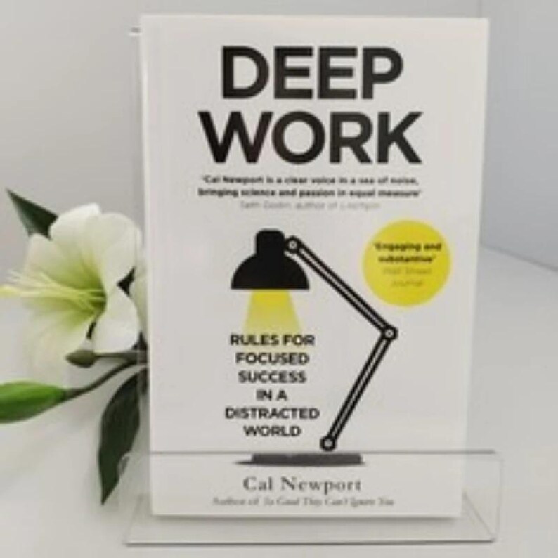 Może przedstawiać: Książka zatytułowana "DEEP WORK" z czarnym tekstem na białym tle. Okładka zawiera czarną grafikę lampy biurkowej i podtytuł "RULES FOR FOCUSED SUCCESS IN A DISTRACTED WORLD" autorstwa Cala Newporta. Biały kwiat i liście znajdują się po lewej stronie.