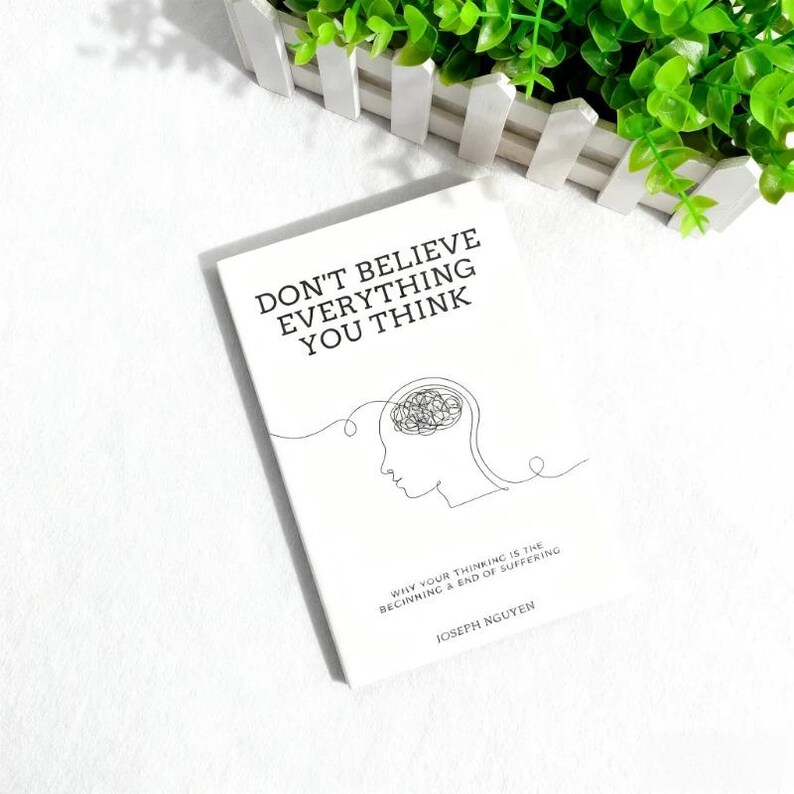 Może przedstawiać: Biała książka zatytułowana "DON'T BELIEVE EVERYTHING YOU THINK" z minimalistyczną ilustracją głowy i m&oacute;zgu. Podtytuł książki brzmi "WHY YOUR THINKING IS THE BEGINNING & END OF SUFFERING" autorstwa Josepha Nguyena.