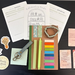 May include: An assortment of stationery and accessories, including a Bible reading tracker, journal, stickers, a keychain, pens, sticky notes, and a bracelet. The items are in shades of green, brown, and white, with floral and inspirational designs.