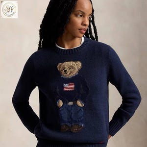 Puede incluir: Suéter azul marino con un diseño detallado de oso que lleva una camisa azul marino, vaqueros azules y zapatos marrones. El oso tiene una bandera americana en su camisa. El suéter tiene un dobladillo y puños acanalados.