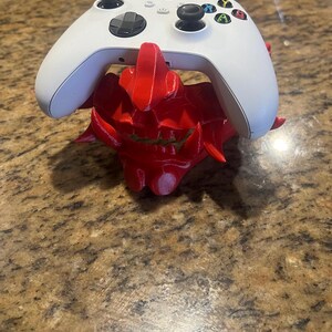 May include: A white Xbox controller rests on a vibrant red, 3D-printed gaming controller stand. The stand has a detailed, spiky design. The controller features black joysticks and colored action buttons.