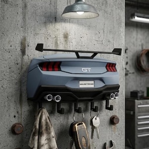 Peut inclure: Un porte-clés mural en forme de l'arrière d'une Ford Mustang GT grise, avec un aileron noir, des feux arrière rouges et quatre pots d'échappement. La plaque d'immatriculation indique MUSTANG, et cinq crochets noirs portent des clés.