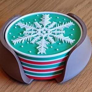 Op de afbeelding: Een ronde onderzetter met een wit sneeuwvlokontwerp op een groene achtergrond, met horizontale rode, witte en groene strepen aan de zijkant, geplaatst in een gebogen, matbruine houder die de onderzetter op zijn plek houdt.