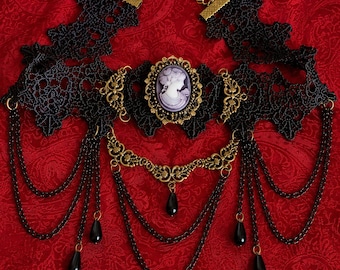 Op de afbeelding: Een zwarte kanten choker met een ovaalvormige camee-hanger met een wit profielsilhouet op een paarse achtergrond. Het sieraad is voorzien van antiek goudkleurig metaalfiligraan, meerdere zwarte kettingen en bungelende zwarte druppelvormige kralen.