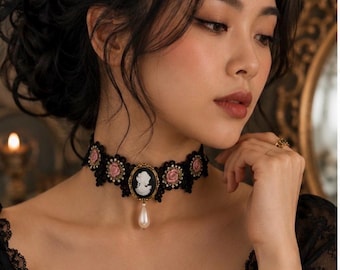 Op de afbeelding: Een zwarte kanten choker met een centraal ovaal camee-hangertje met een wit profielsilhouet op een zwarte achtergrond, geaccentueerd door een bungelende witte druppelvormige parel en kleine roze roosjes langs de kanten band.