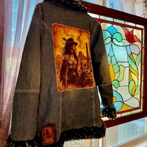 May include: A blue denim jacket featuring a large rectangular patch on the back depicting a person in a hat standing in a desert landscape with cacti. The jacket has dark faux fur trim along the collar, cuffs, and hem, and a small rectangular patch.