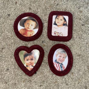 May include: Four dark red velvet-textured picture frames in various shapes including a circle, a rectangle with rounded corners, a heart, and an oval. Each frame contains a photograph of a child, displaying different facial expressions and clothing.
