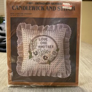 May include: A packaged Candlewick and Stitch pillow kit. The pillow features a floral design with the words "LOVE ONE ANOTHER" embroidered in a circle. The pillow measures 14 x 14 inches and has a lace trim.