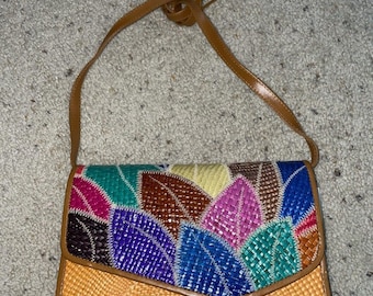 Peut inclure: Un sac à bandoulière tressé rectangulaire avec une base beige et un rabat orné d'un motif de feuilles multicolores dans des tons de bleu, vert, rose, jaune et marron. Le sac comprend une fine bandoulière assortie de couleur beige.