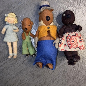 May include: Four vintage felt dolls. A blonde girl in a blue dress, a brown bear in green overalls, a bear in a hat, yellow vest, and blue pants, and a black doll in a floral skirt.