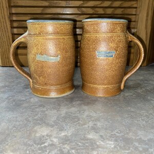May include: Two brown ceramic mugs with handles. One mug features a canoe design, the other has the text "WATERLAWN COTTAGES" in a blue rectangle. Both mugs have a light blue rim.