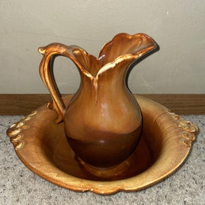 Op de afbeelding: Een waskom en bijpassende kan van keramiek met een glanzend, tweekleurig bruin glazuur. De kan heeft een gebogen handvat en een geschulpte rand, terwijl de kom een brede, ronde vorm heeft met decoratieve, gevormde randen aan beide zijden.