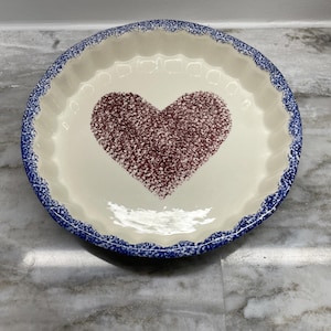 May include: A round ceramic pie dish with a cream-colored interior featuring a large, dark red speckled heart design in the center. The rim of the dish has a scalloped edge and is decorated with a blue and white speckled sponge-painted pattern.