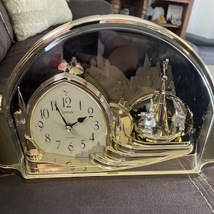 May include: A gold-toned quartz clock with a cream-colored face and black numerals. The clock features a decorative scene with a castle and a pendulum. The clock is encased in a clear, arched cover.