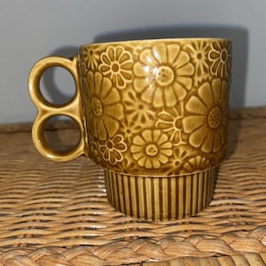 Könnte beinhalten: Eine braune Keramiktasse mit floralem Muster. Die Tasse hat einen einzigartigen Griff mit zwei kreisförmigen Öffnungen. Der Boden der Tasse hat vertikale Linien. Die Tasse steht auf einer gewebten Oberfläche.