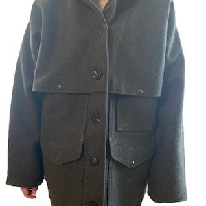 Op de afbeelding: Donkergroene wollen jas met een cape-achtige overlay, met een kraag en knoopsluiting. De jas heeft twee grote voorzakken en een lichtere bruine voering zichtbaar bij de kraag. Een klassiek bovenkledingstuk.