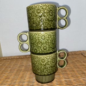 May include: Three stacked green ceramic mugs with a floral pattern. Each mug has a unique handle with two circular openings. The mugs are a muted green color and are stacked on top of each other.