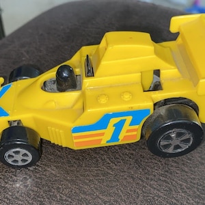 May include: Yellow toy race car with black wheels and a rear spoiler. The car has blue and orange stripes with the number 1 on the side. A small black figure sits in the cockpit.