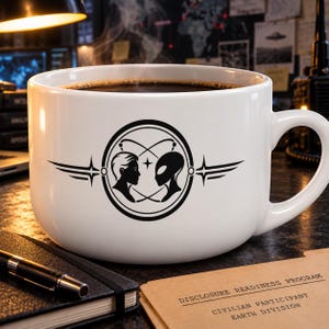 Può includere: Una tazza in ceramica bianca con una grafica nera che mostra un profilo umano di fronte a un profilo alieno all'interno di un emblema circolare con ali stilizzate. La tazza contiene un liquido scuro ed è posizionata su una scrivania.