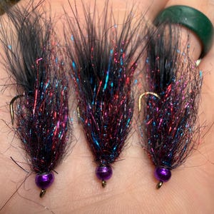 May include: Three fishing flies with black, purple, red, and blue detailing. Each fly has a dark purple bead and a gold hook. These flies are designed for angling.