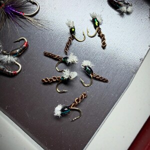 May include: Close-up of several handmade fishing flies. The flies feature gold hooks, brown twisted bodies, and white and green accents. Other flies in the image have red, purple, and black details. The flies are arranged on a dark surface.