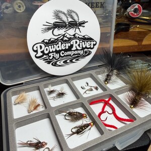 May include: A clear plastic fly fishing tackle box with a white Powder River Fly Company sticker. The box contains various artificial flies in a gray foam insert, including red, brown, and black designs. The flies are for fishing.