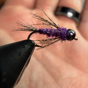 Può includere: Primo piano di un'esca da pesca a mosca viola e oro. L'esca ha una testa nera, un corpo viola e un avvolgimento in filo d'oro. L'esca è tenuta in un attrezzo nero.