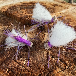 May include: Three fishing flies featuring bright purple bodies, white synthetic hair wings, and thin purple-and-black speckled rubber legs. Each fly is constructed with a metal hook and a small tan foam head, designed for fly fishing applications.