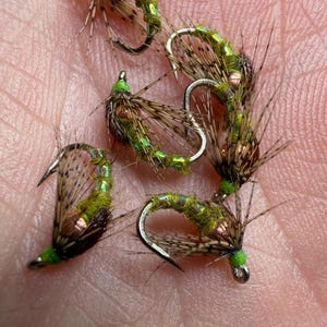 May include: Close-up of several artificial fishing flies. The flies feature a combination of green, brown, and copper-colored materials, with curved hooks. The flies are designed to mimic insects and are used for fly fishing.