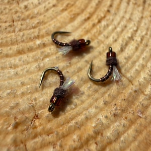 May include: Three fishing flies with curved hooks, brown bodies, and iridescent wings are arranged on a light brown, textured wooden surface. The flies have a dark brown, fuzzy material near the hook bend and a small, gold-colored bead.
