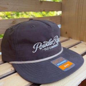 May include: A dark grey baseball cap featuring the embroidered white text Powder River FLY COMPANY. The cap includes a decorative beige rope detail across the base of the crown and a rectangular orange and blue sticker on the flat brim.