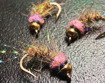 May include: Three handmade fishing flies with gold-colored hooks and heads. The flies have brown and tan bodies with pink and iridescent accents. The flies are designed for fly fishing.