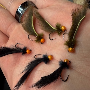 Puede incluir: Primer plano de varias moscas de pesca. Las moscas tienen cuerpos negros o verde oliva parecidos a pieles, cabezas de cuentas naranjas y anzuelos curvos. Las moscas están dispuestas en la palma abierta de una persona.